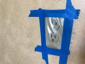 Wall outlet covered with plastic sheeting and painter’s tape.